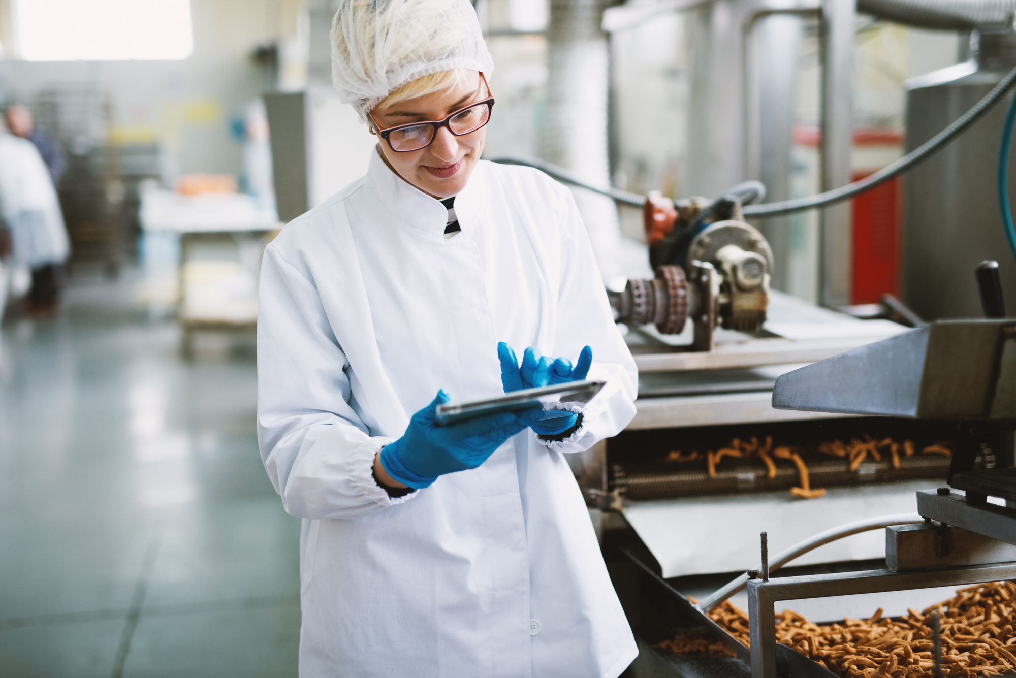 Food Sanitation Worker Reviewing Report On Tablet Next To Conveyor of Pretzels